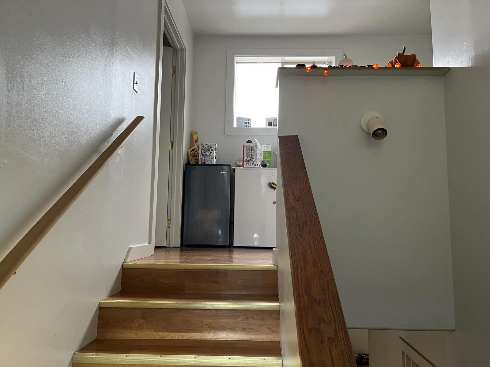 Top of wooden stairs leading to a landing with two compact refrigerators, storage furniture and items near a window.