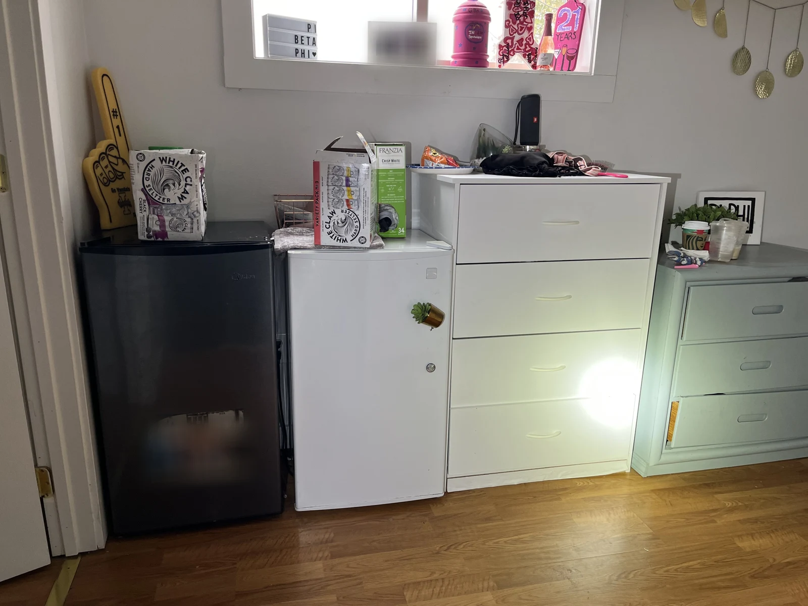 Two compact refrigerators beside white and pale-green drawer units, with drink boxes and other belongings on top.