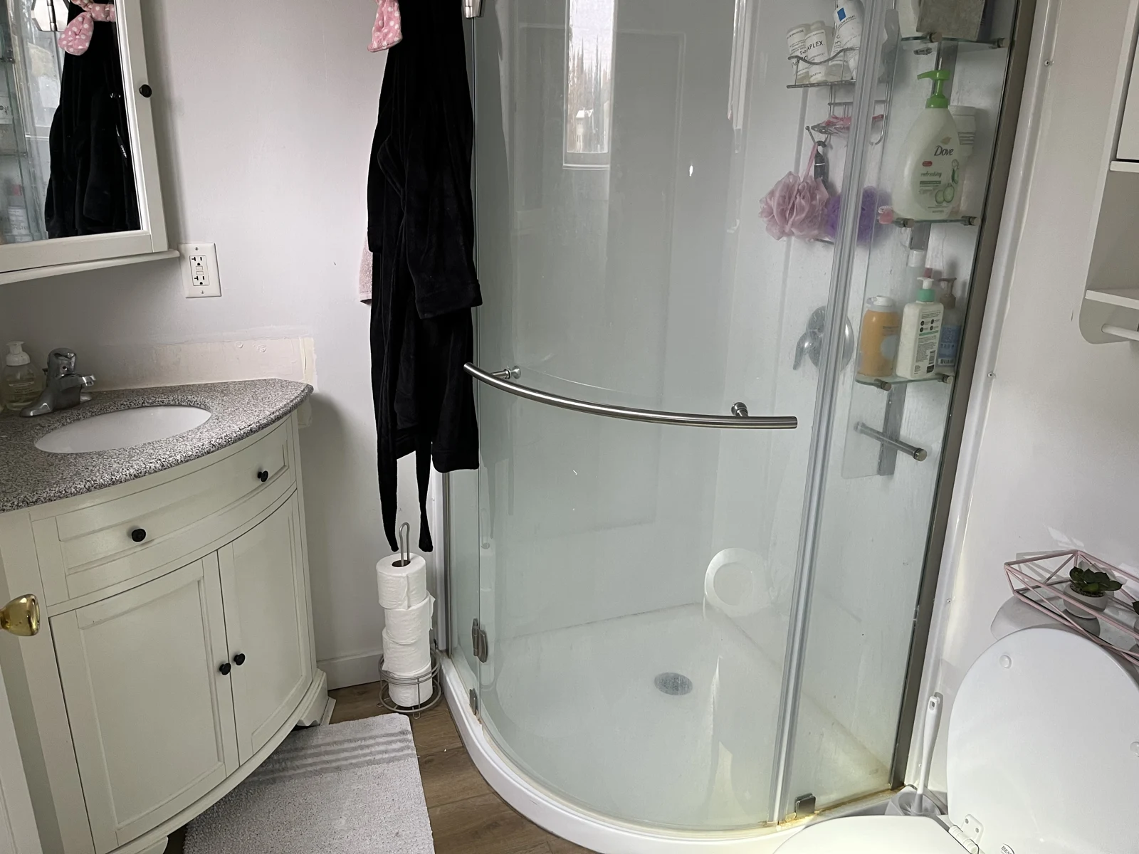 Bathroom with a curved sink vanity, rounded glass shower enclosure, toiletries, hanging dark garment and stacked toilet-paper rolls.