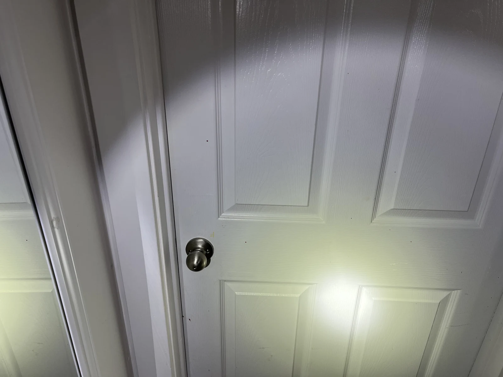 Closed white paneled door with a round metal knob, lit by a flashlight.