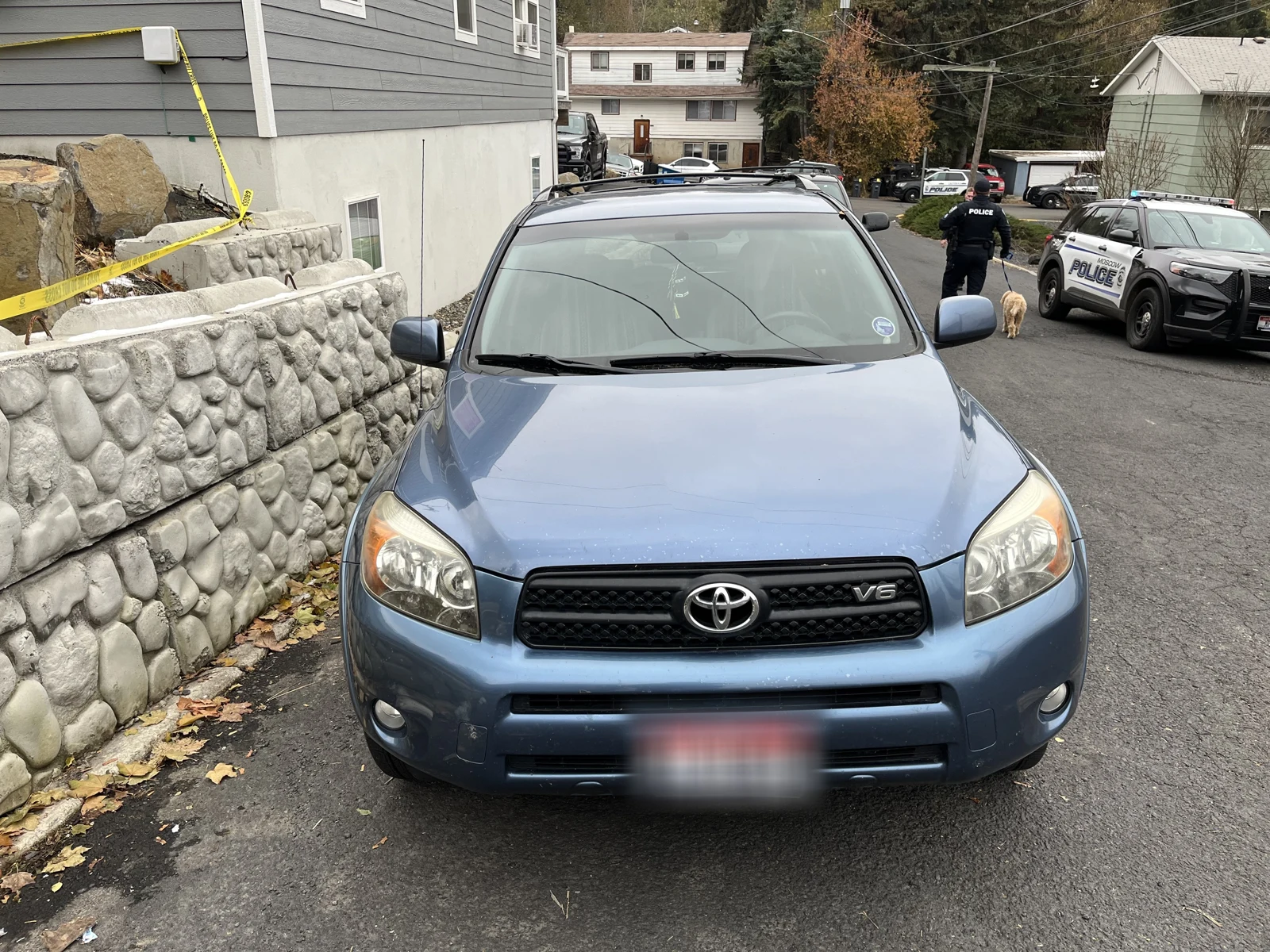 Front of a blue Toyota SUV beside a stone retaining wall, with a blurred plate and police vehicle in the street behind.