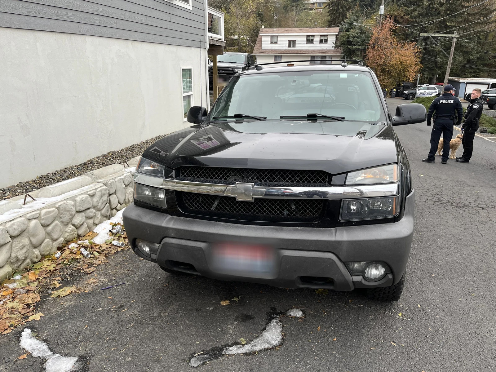 Front of a black Chevrolet SUV with a blurred plate, beside a retaining wall and people standing farther along the street.