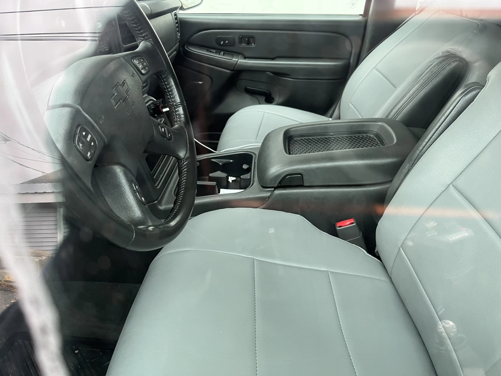 Light-gray leather front seats, Chevrolet steering wheel and center armrest with a mesh tray, viewed through vehicle glass.