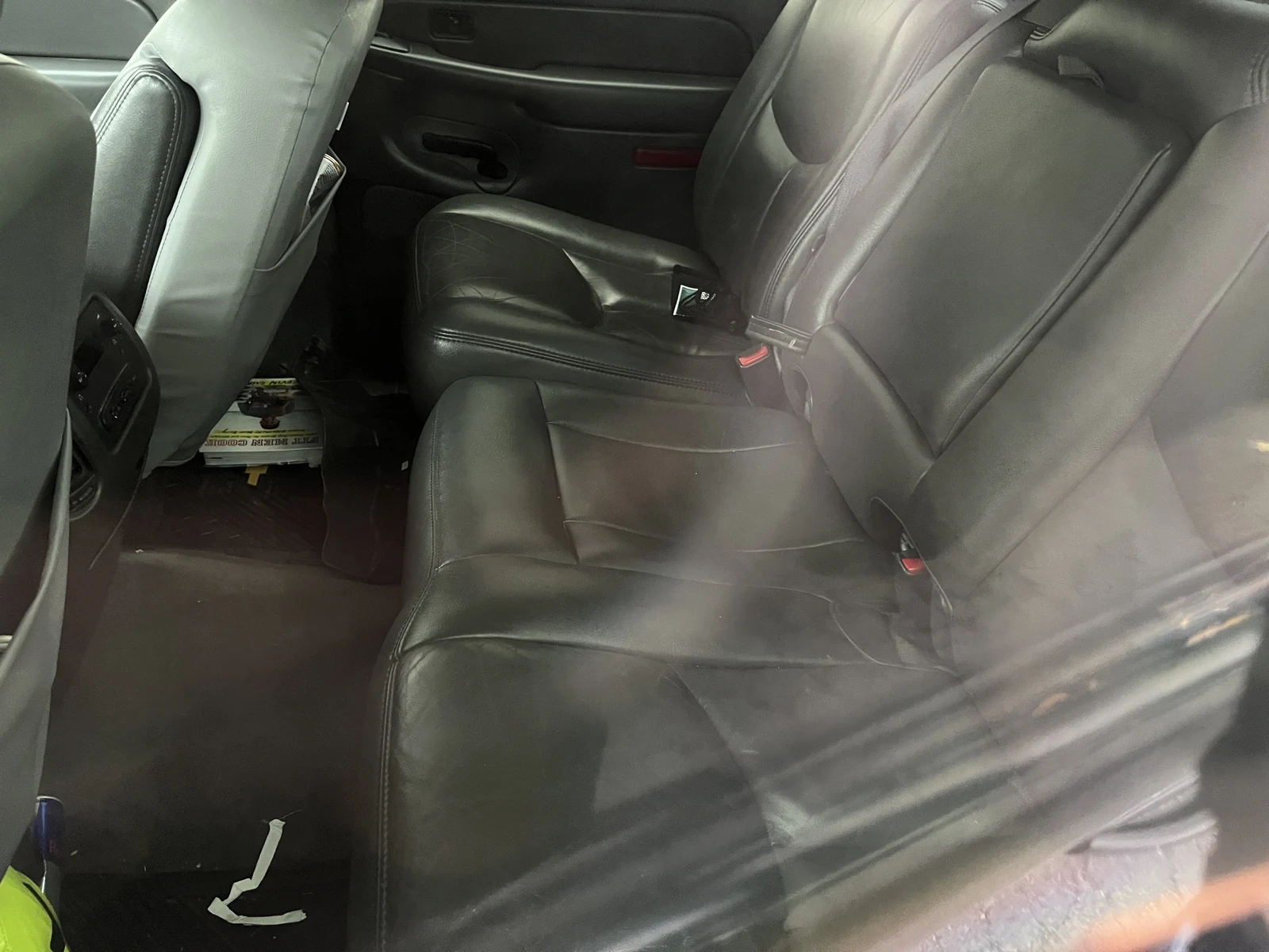 Gray leather rear bench seat with seat-belt buckles and small items on the floor, viewed through glass.