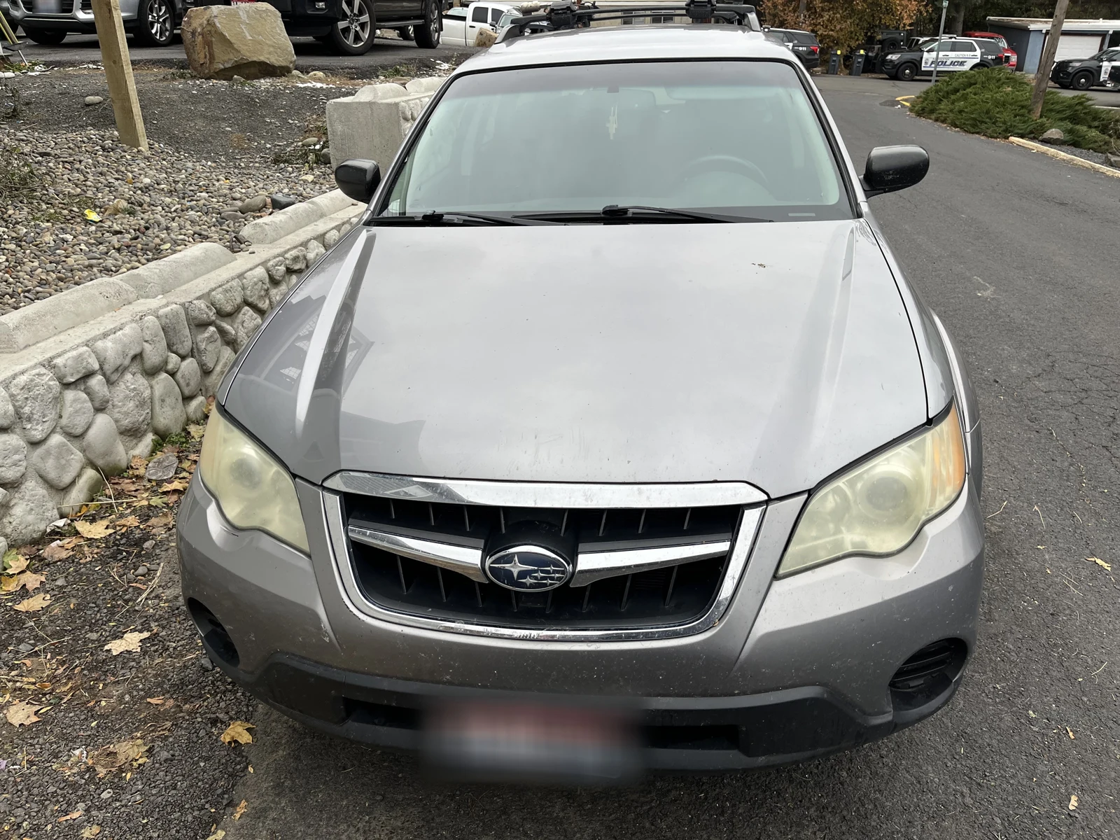 Front of a gray Subaru with roof rails and a blurred plate, parked beside a stone retaining wall.