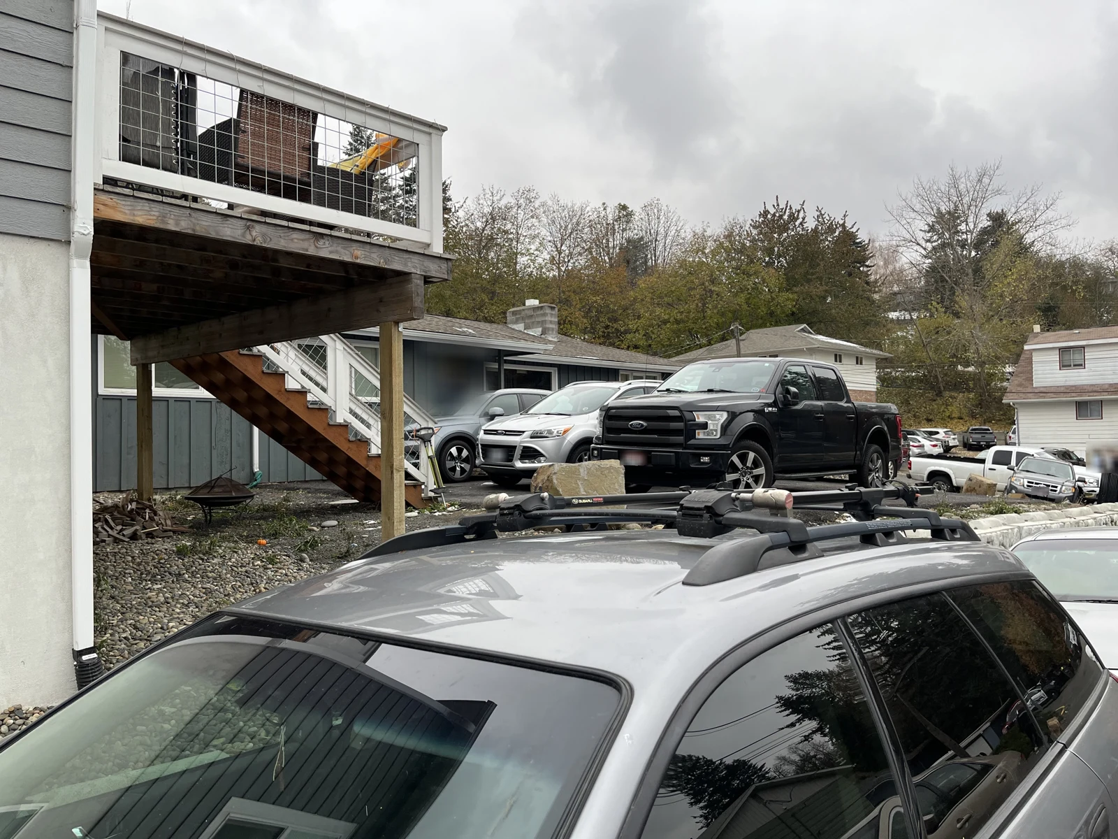 Upper side and roof rack of a gray station wagon, with a wooden exterior staircase and parked vehicles behind.