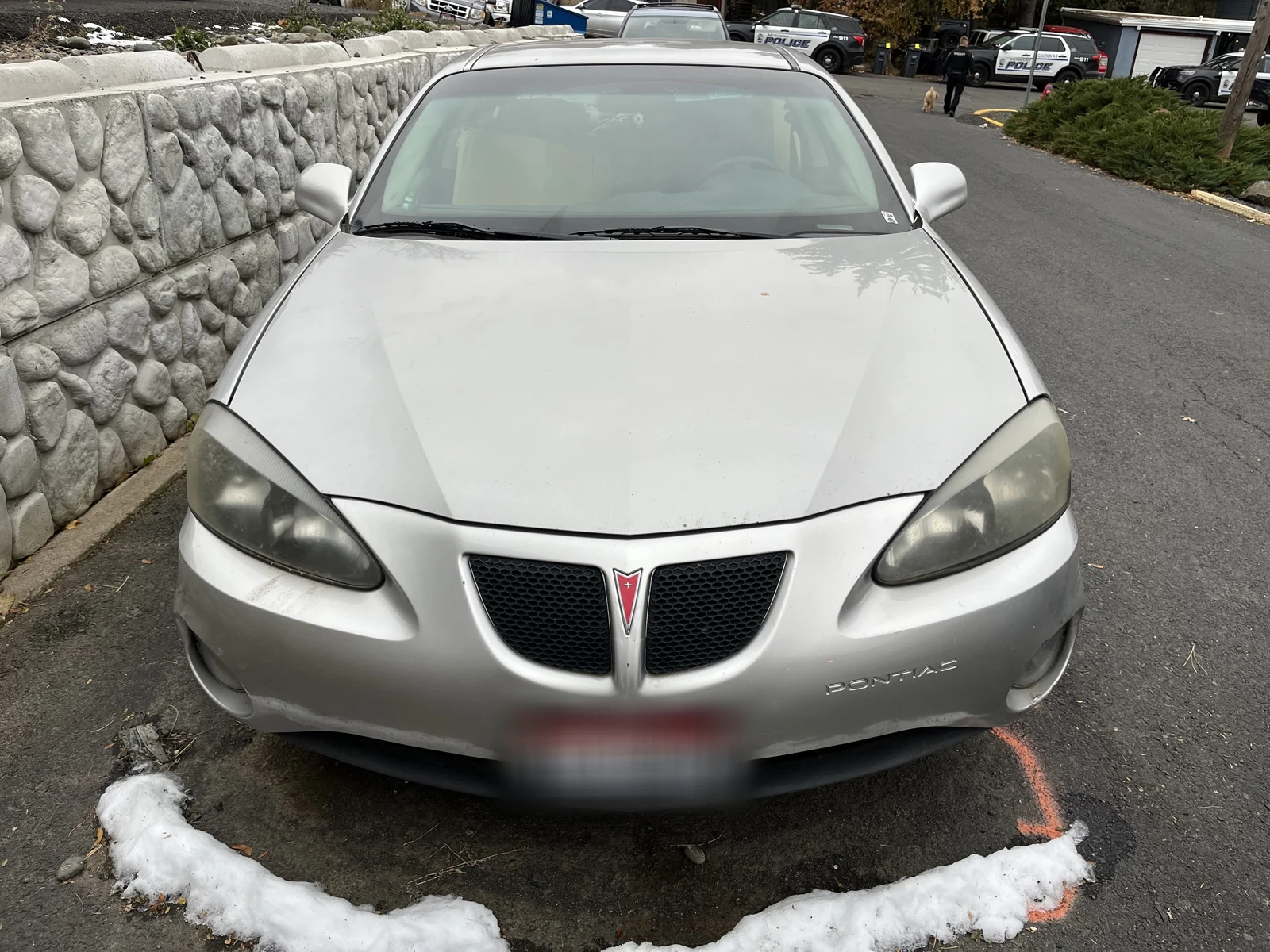 Front of a silver Pontiac Grand Prix with a blurred plate and a patch of snow on the ground.