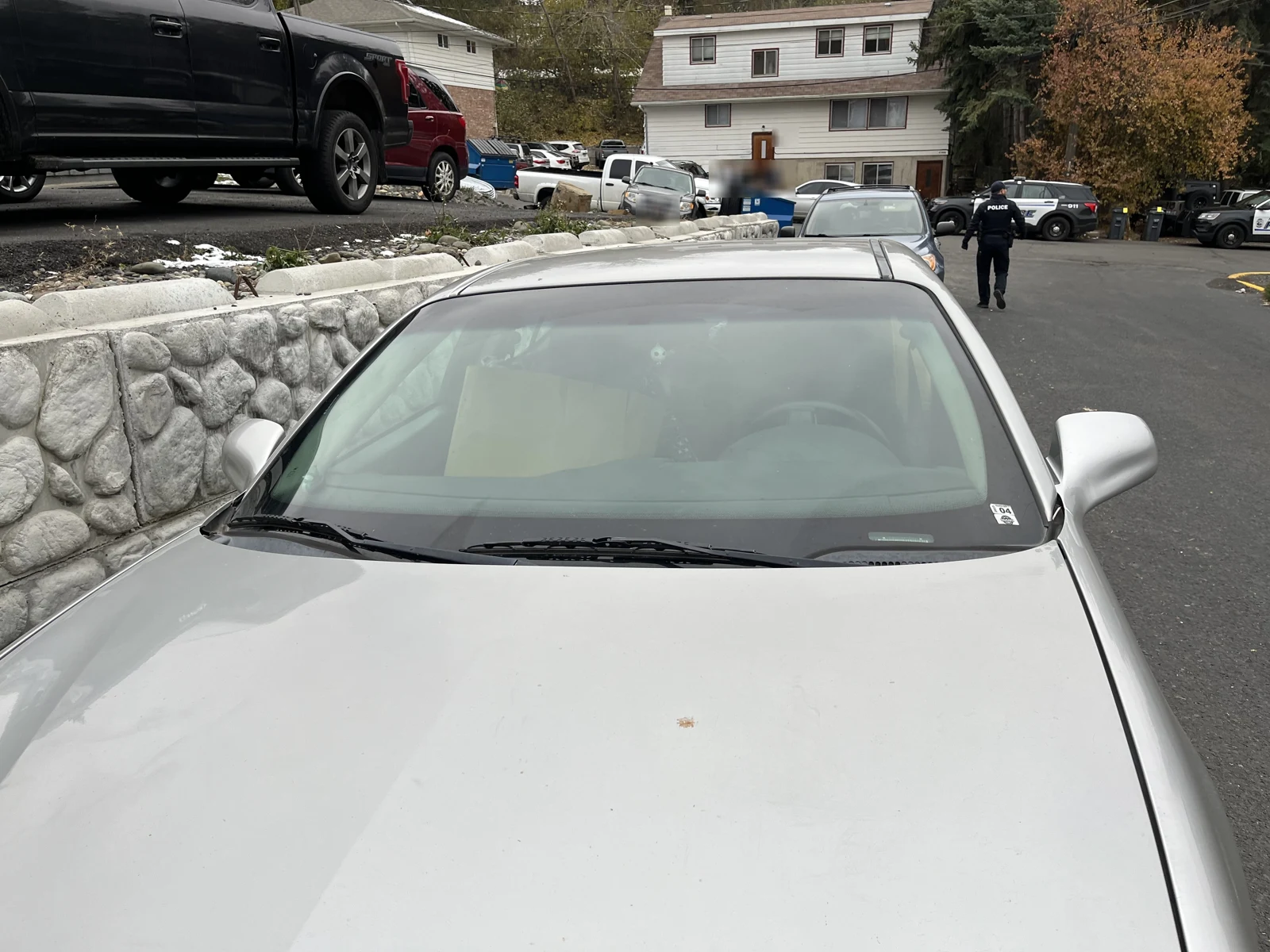 Hood and windshield of a silver car beside a stone wall, with parked vehicles along the street beyond.