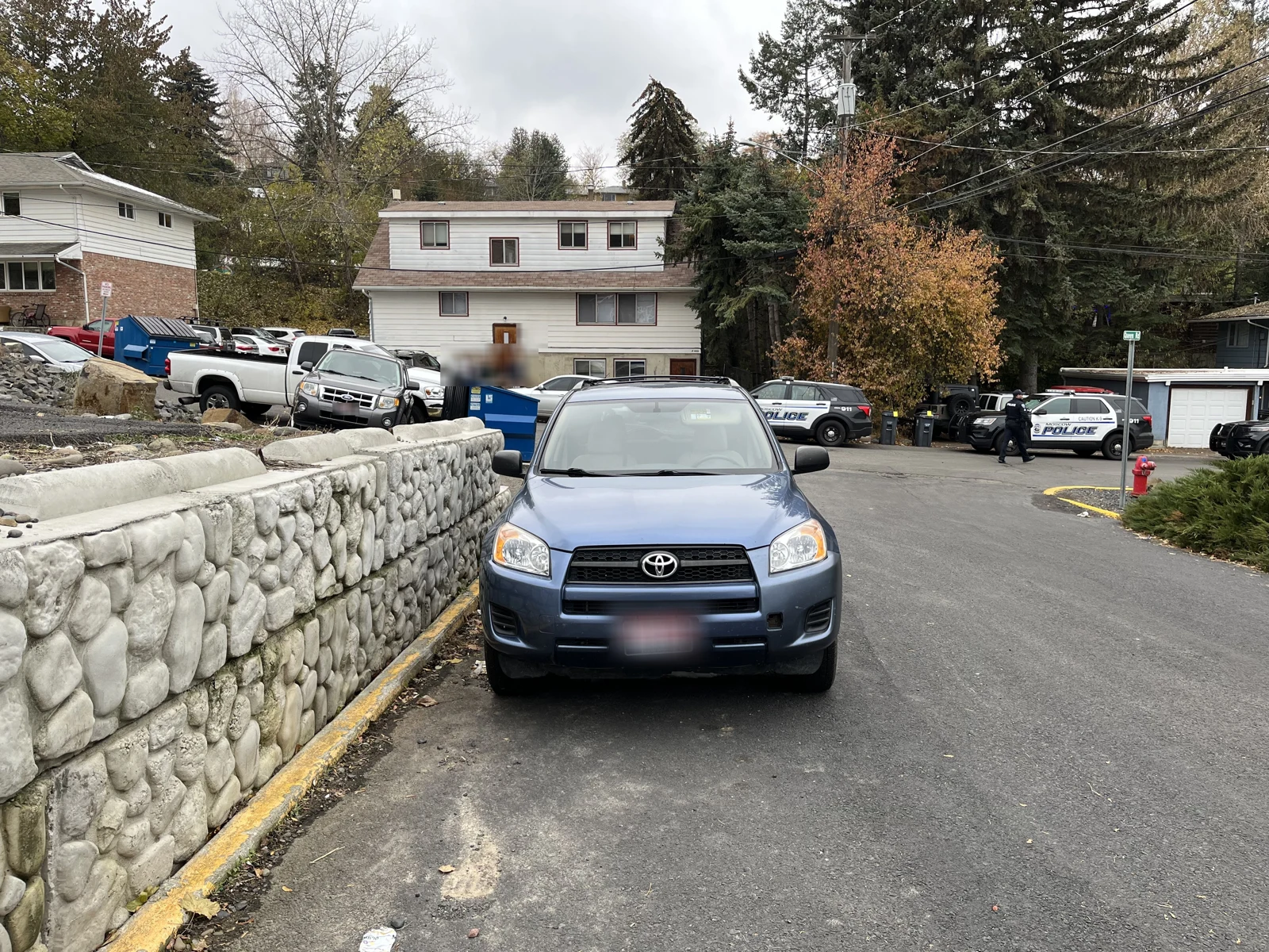 Blue Toyota SUV parked along a stone retaining wall, with houses, other vehicles and a police car farther away.