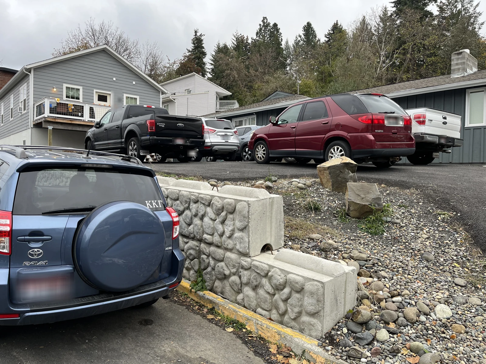 Rear of a blue Toyota SUV below a retaining wall, with a maroon minivan and pickup trucks parked on higher ground.