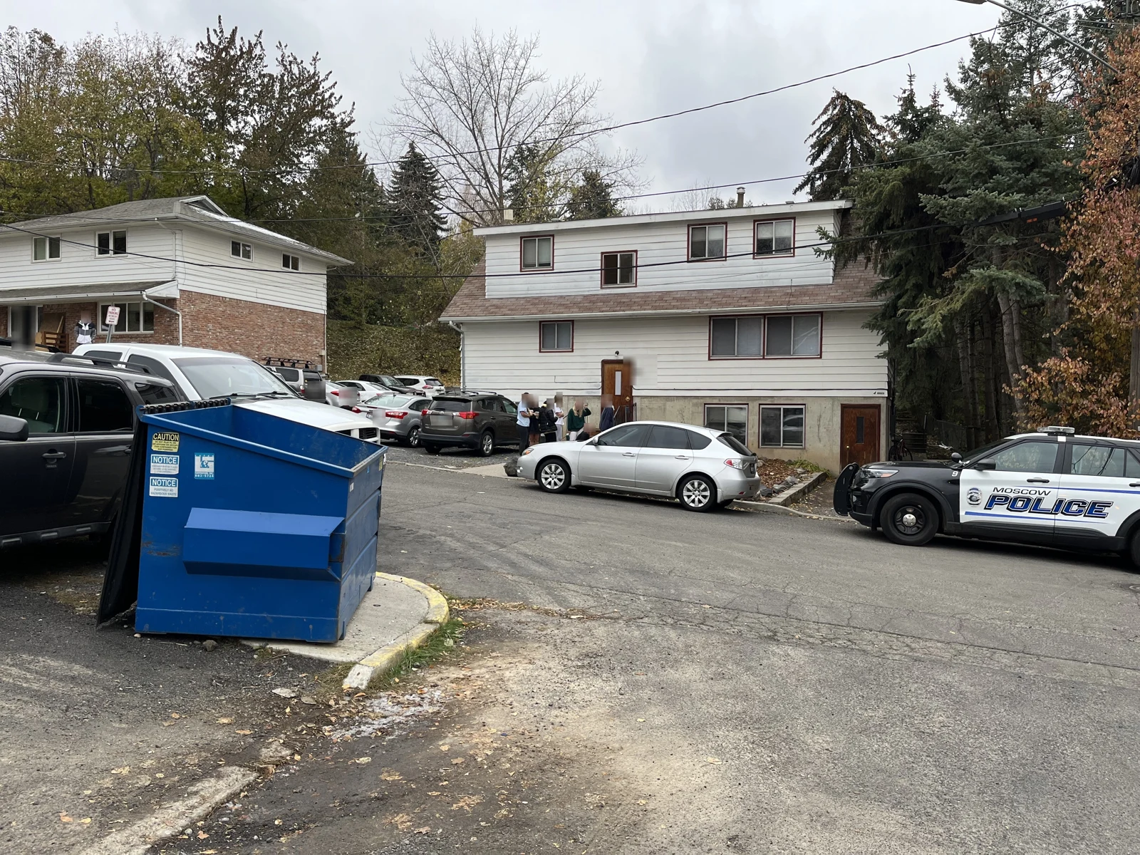 Blue dumpster and parked cars on a residential street, with two-story houses and a marked police vehicle.
