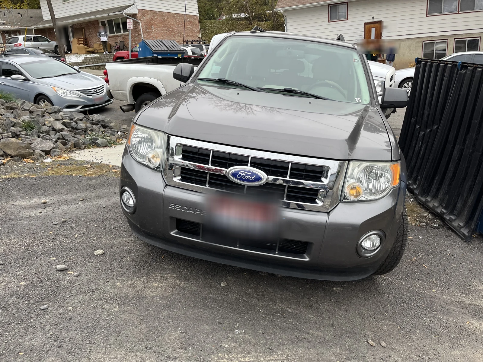 Front of a gray Ford pickup with a blurred plate, parked beside a dark fence and a rocky slope.