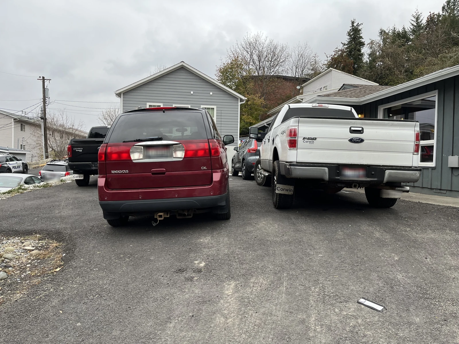 Rear of a maroon minivan and a white Ford pickup parked in a driveway beside gray buildings.