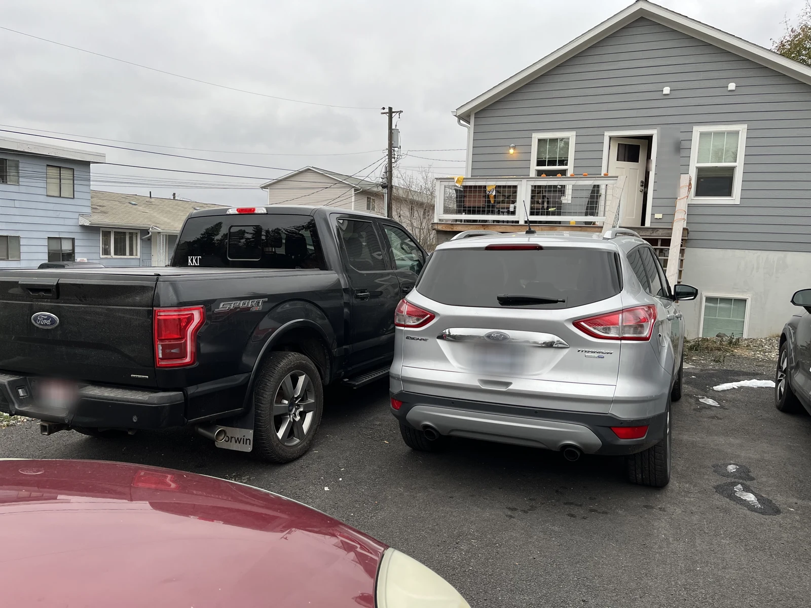 Rear of a black Ford pickup and silver SUV parked outside a gray house with a small raised porch.