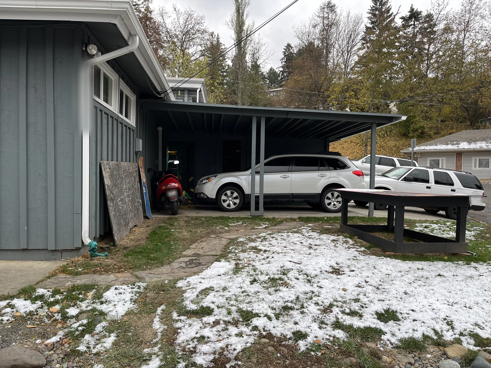 House side and carport sheltering a white SUV, with a picnic table and snow on the lawn.