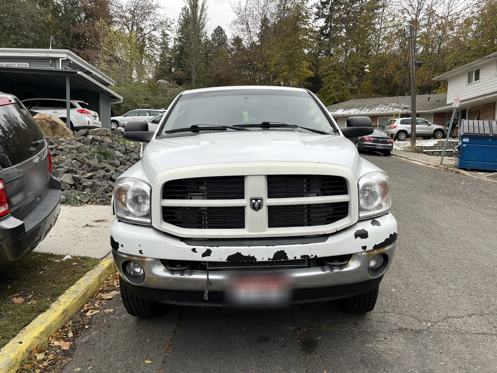 Front of a white Dodge pickup with peeling dark patches on the bumper and a blurred license plate.