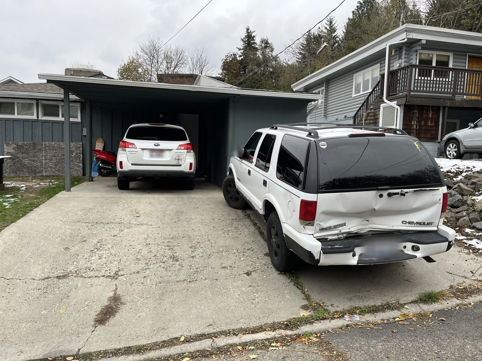 Rear of a white Chevrolet SUV with a dented rear hatch and damaged bumper, beside a driveway and carport.