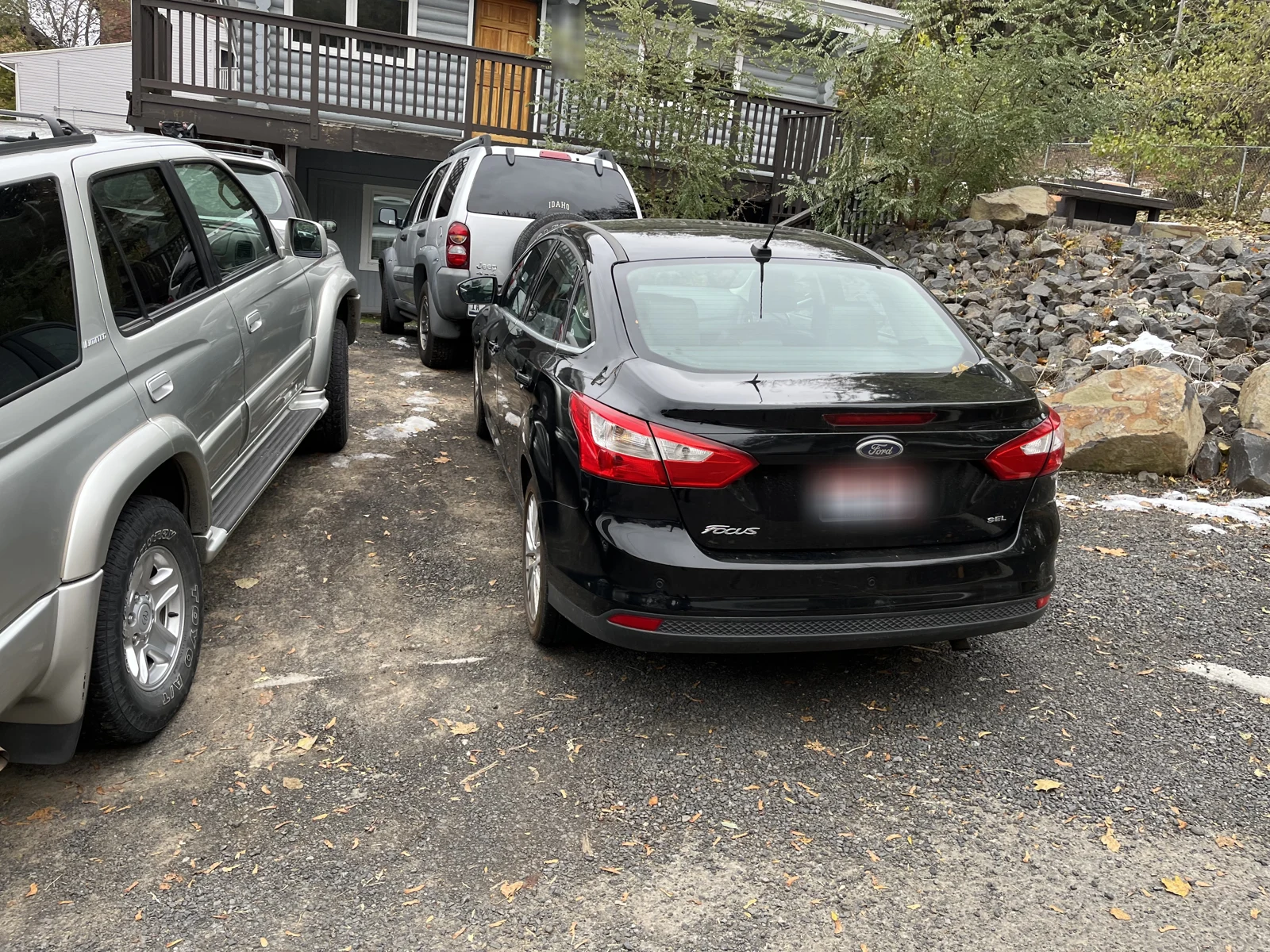 Rear of a black Ford Focus with a blurred plate among parked vehicles beside a rocky slope.