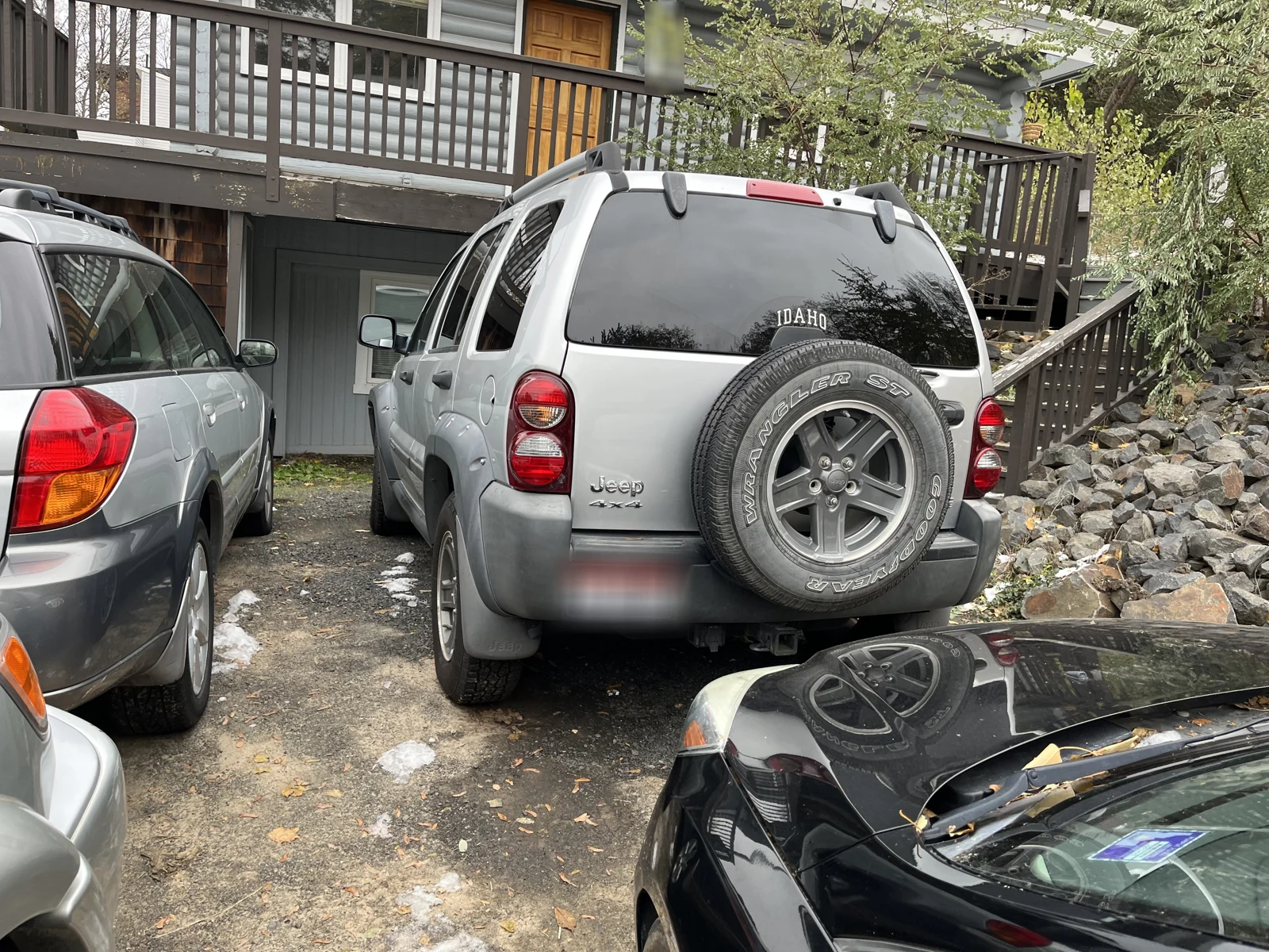 Rear of a silver Jeep with a spare tire, parked among vehicles below a wooden balcony.