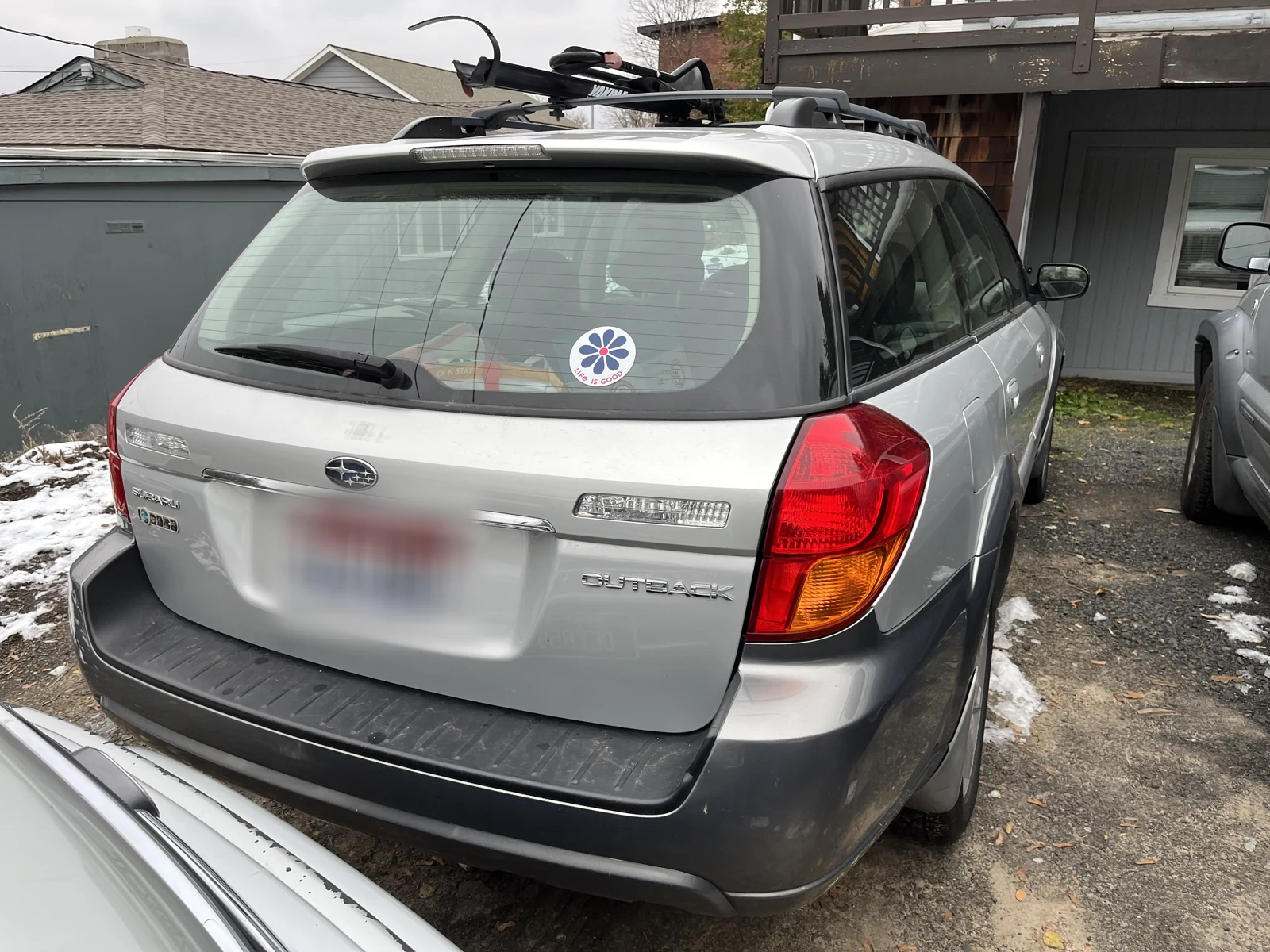 Rear of a silver Subaru Outback with a roof rack, circular rear-window sticker and blurred plate.