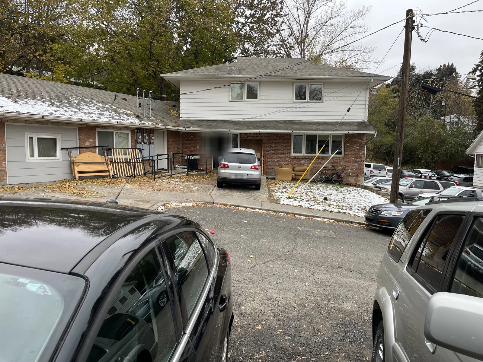 Residential street viewed between parked vehicles, with a two-story house, garage entrance and snow on the ground.