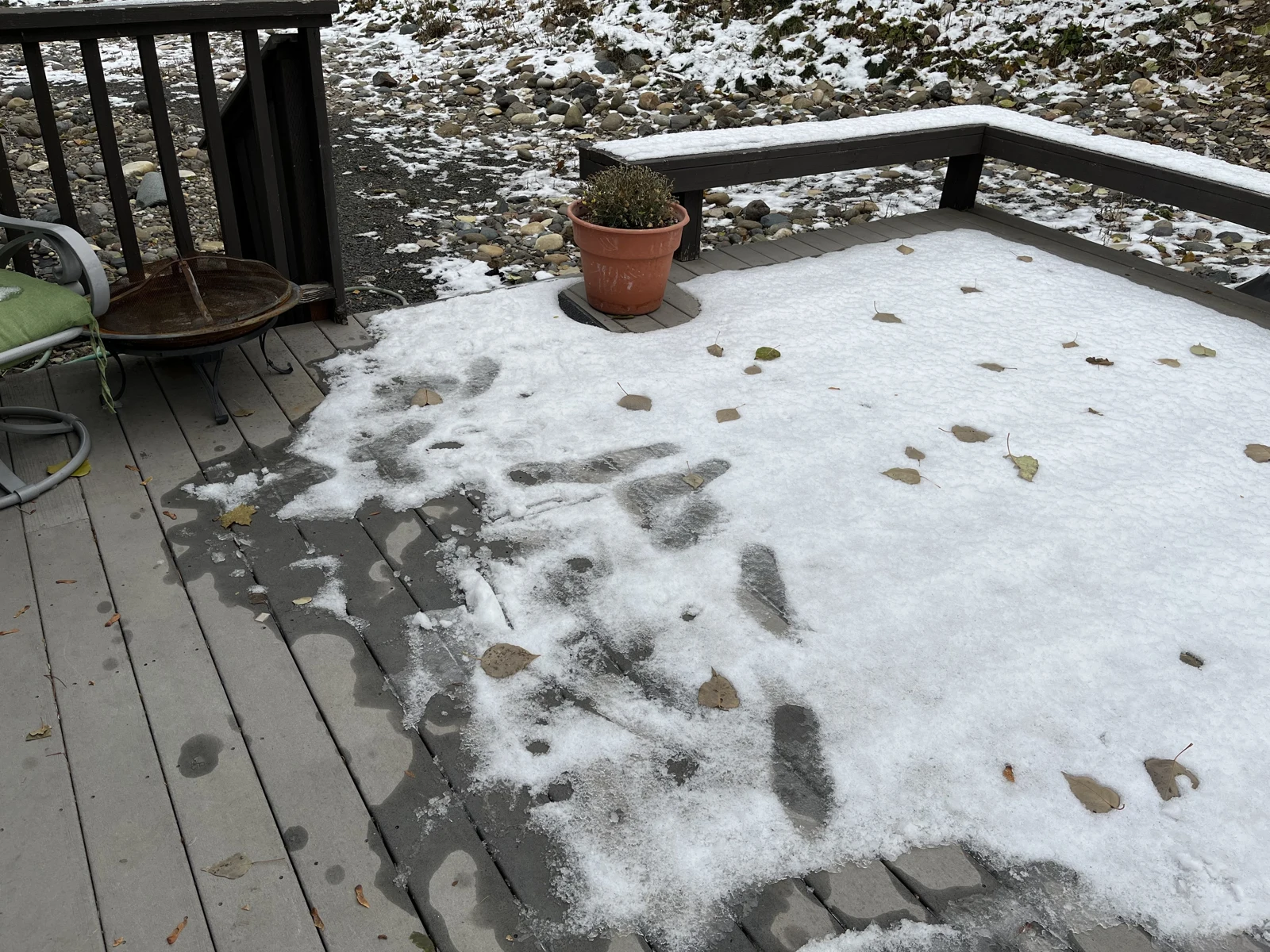 Snow-covered wooden porch with visible shoe-shaped impressions, fallen leaves, a potted plant and railing.