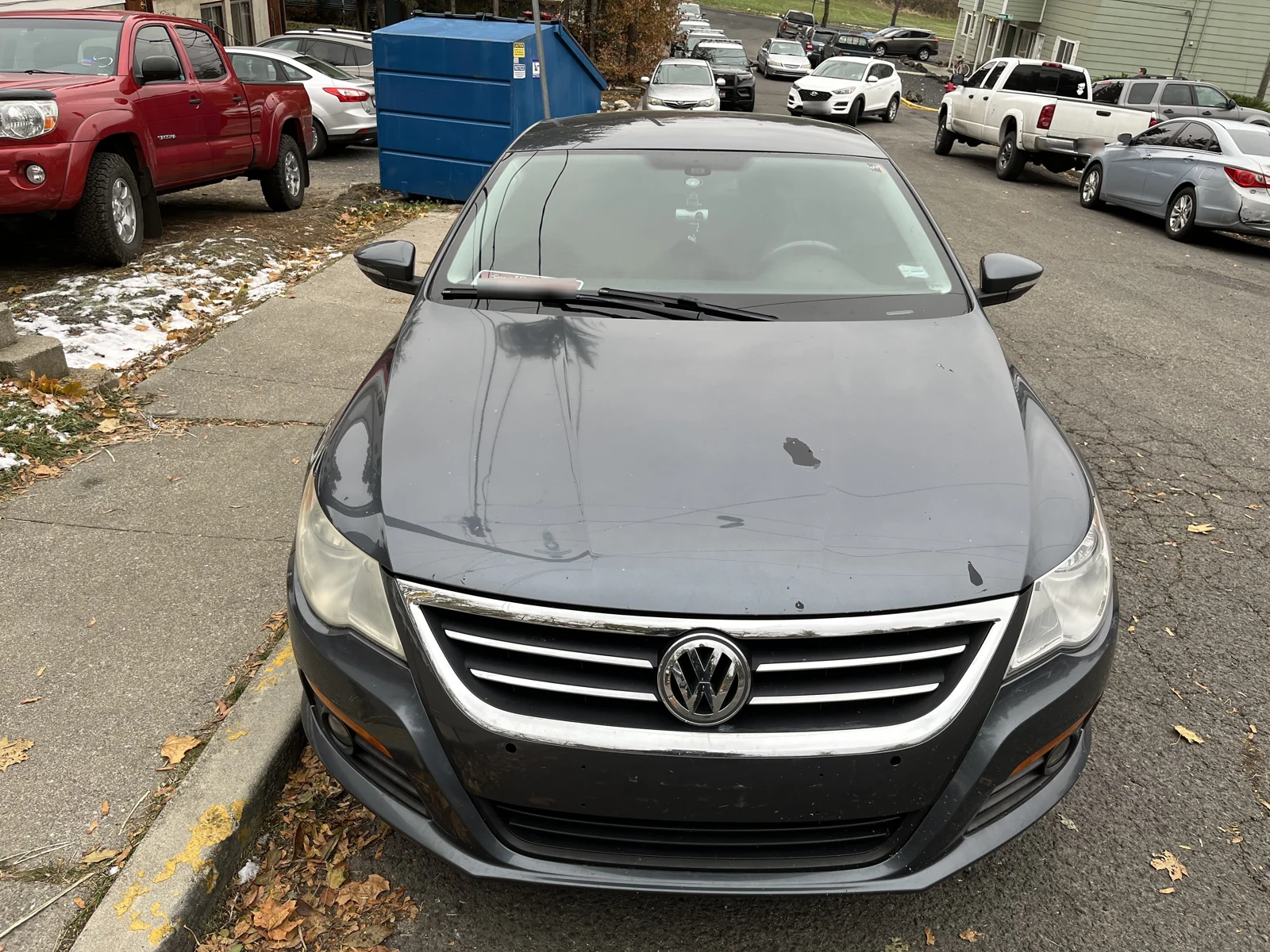 Front of a black Volkswagen sedan parked on a residential street, with a blue dumpster and other vehicles behind.