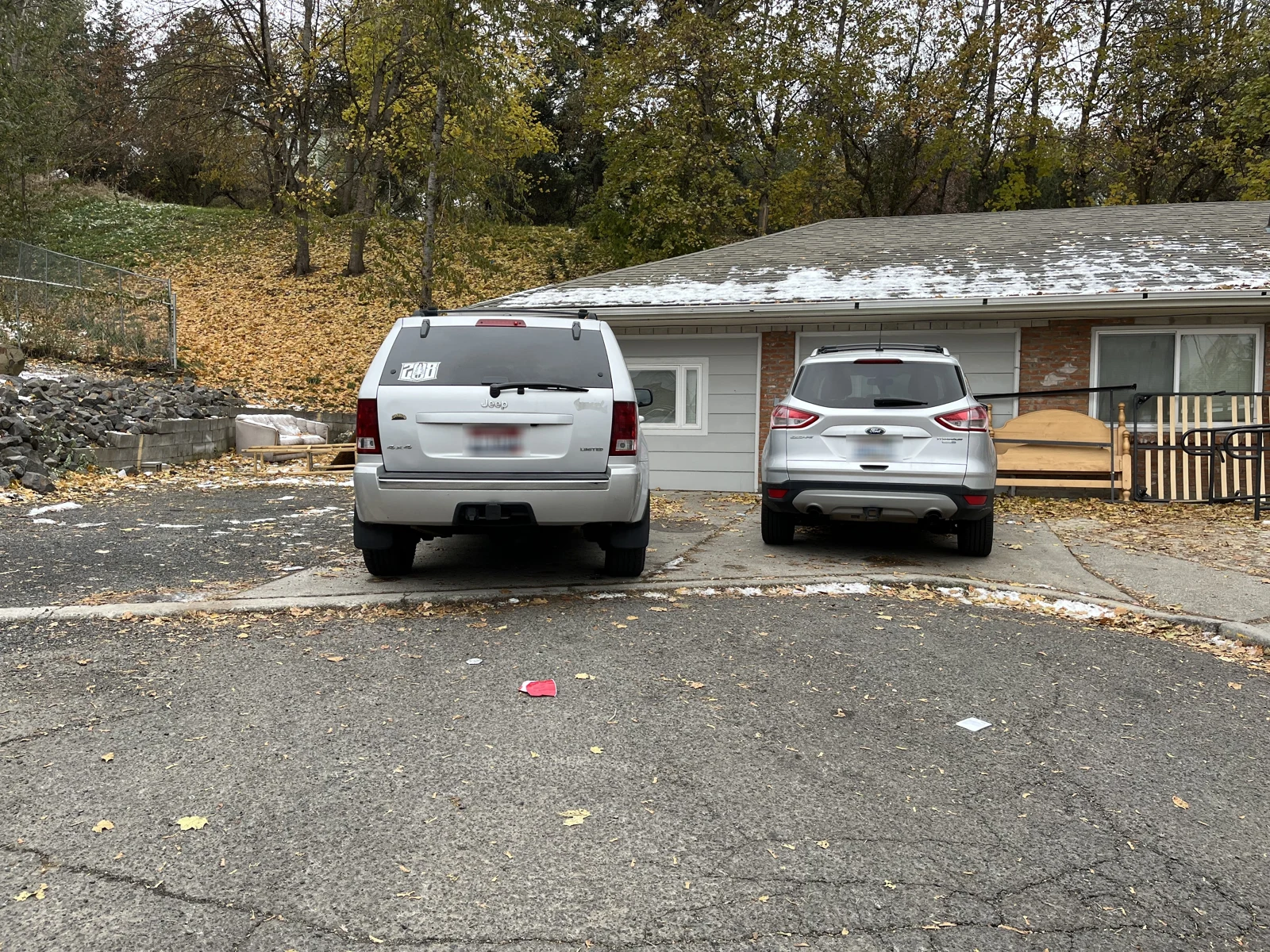 Two light-colored SUVs parked beside a low gray house with snow on the roof and a wooden railing.