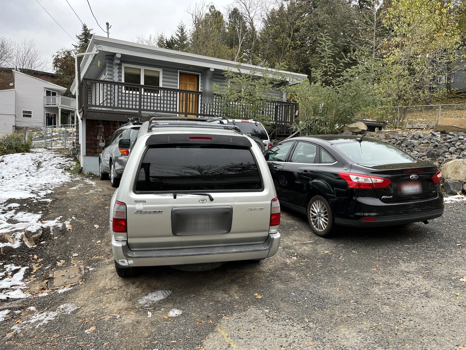 Rear views of a silver SUV and black sedan outside a gray house with a raised balcony and patches of snow.