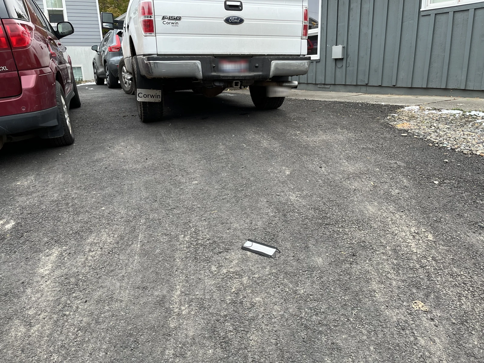 Disposable face mask on asphalt below a white pickup truck and maroon vehicle.