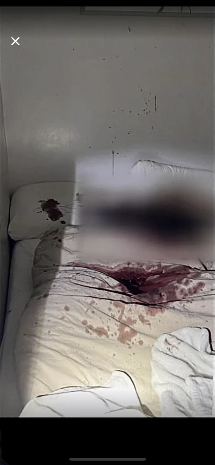 Phone screenshot showing bedding and pillows with dark red stains and a large blurred area, with a white wall behind the bed.