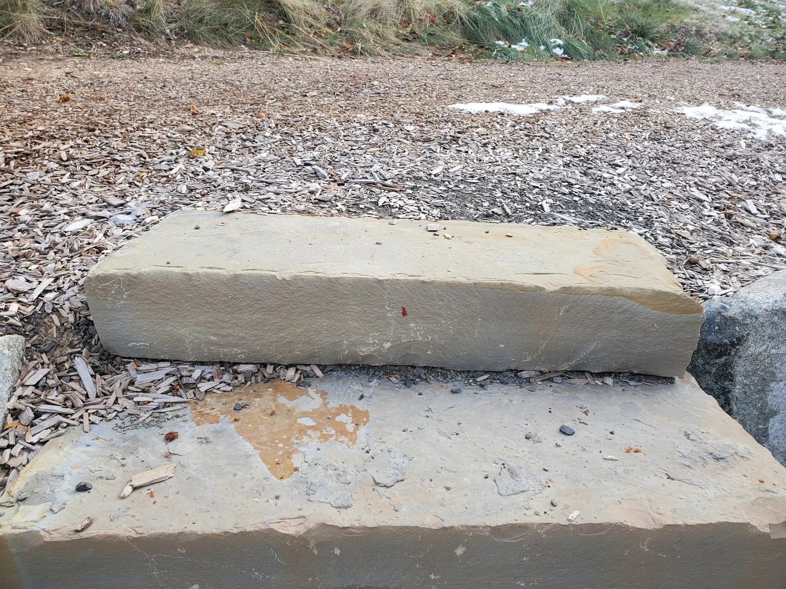 Rectangular stone block with a reddish mark on its face, surrounded by gravel, wood chips and patches of snow.