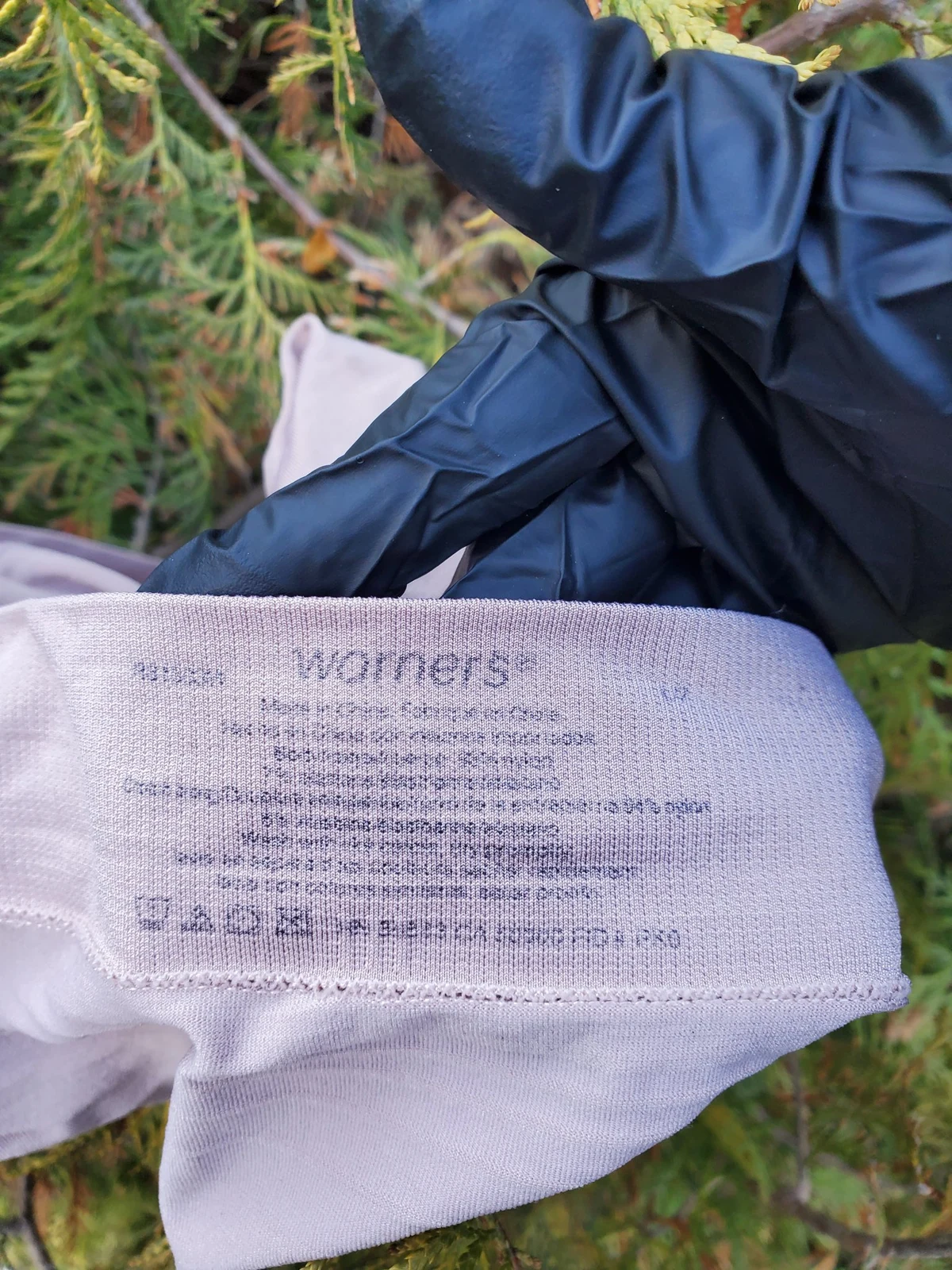 Black-gloved hand holding pale purple underwear with a printed Warners care label, against green branches.