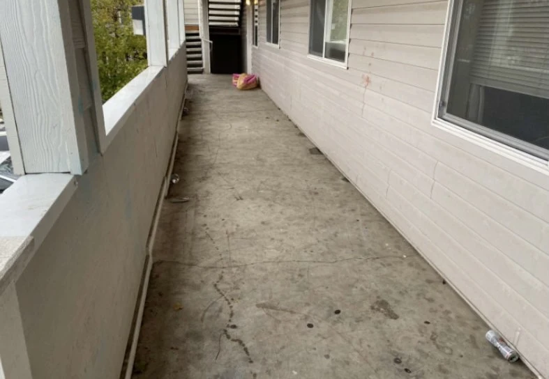 Long exterior apartment walkway with windows, pale siding, a concrete floor and stairs at the far end.