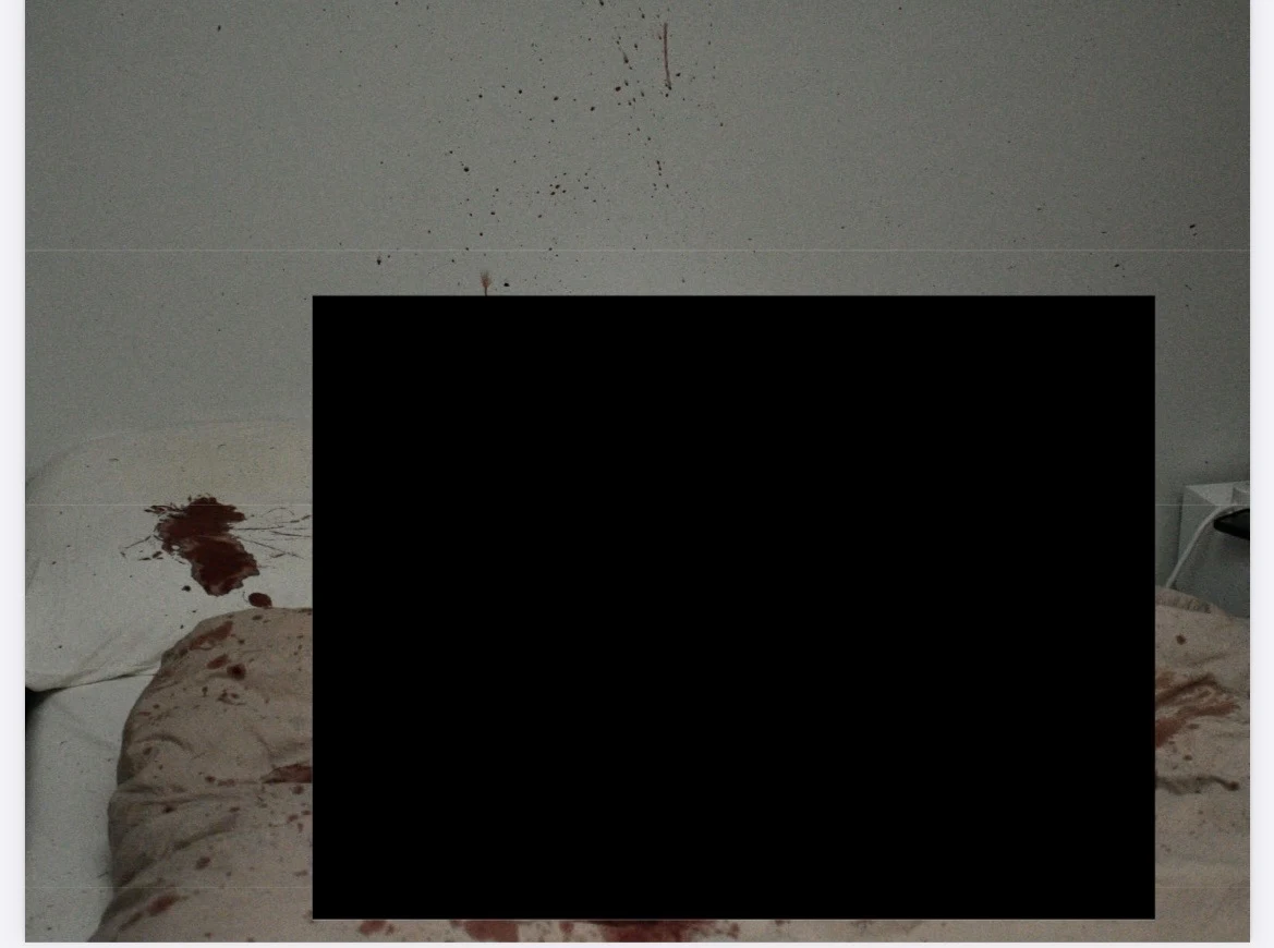 Bed, pillow and wall with dark reddish stains, largely obscured by a solid black redaction.