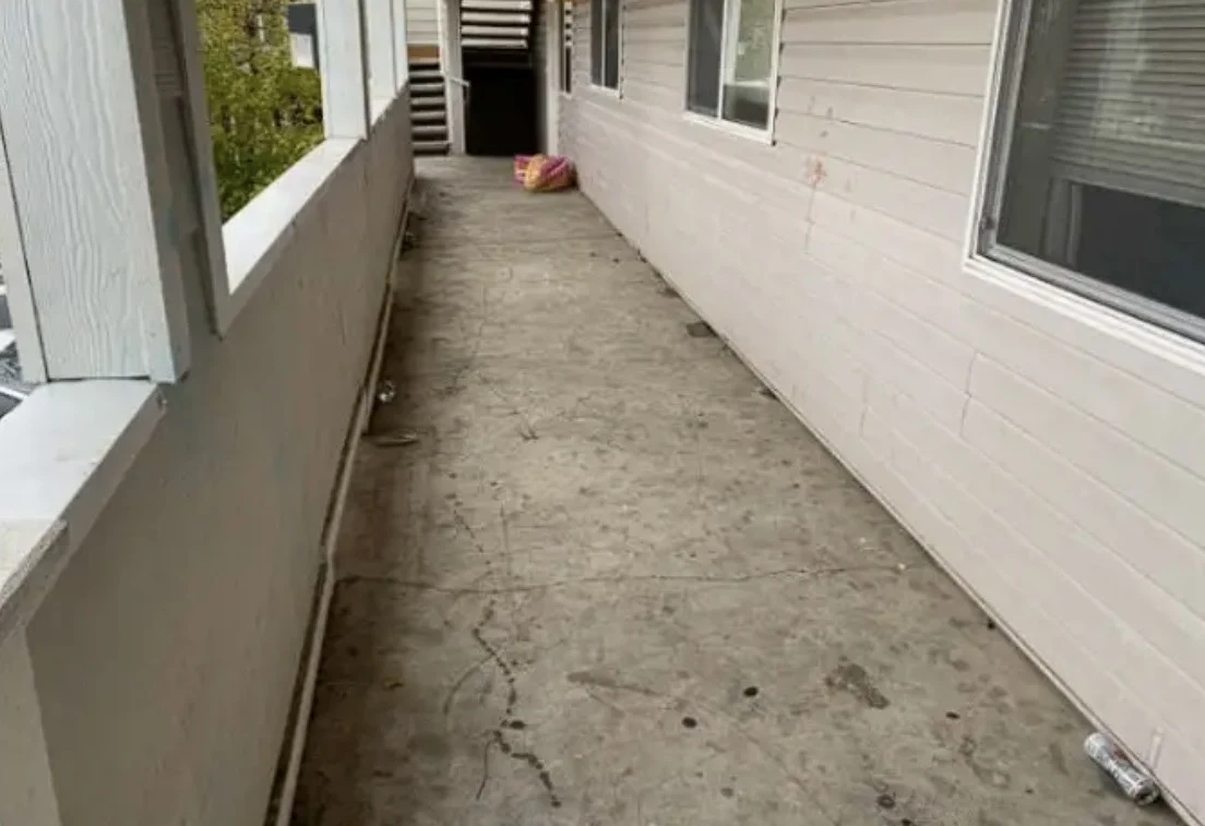 Exterior apartment walkway with pale siding, windows, concrete floor and stairs at the far end.
