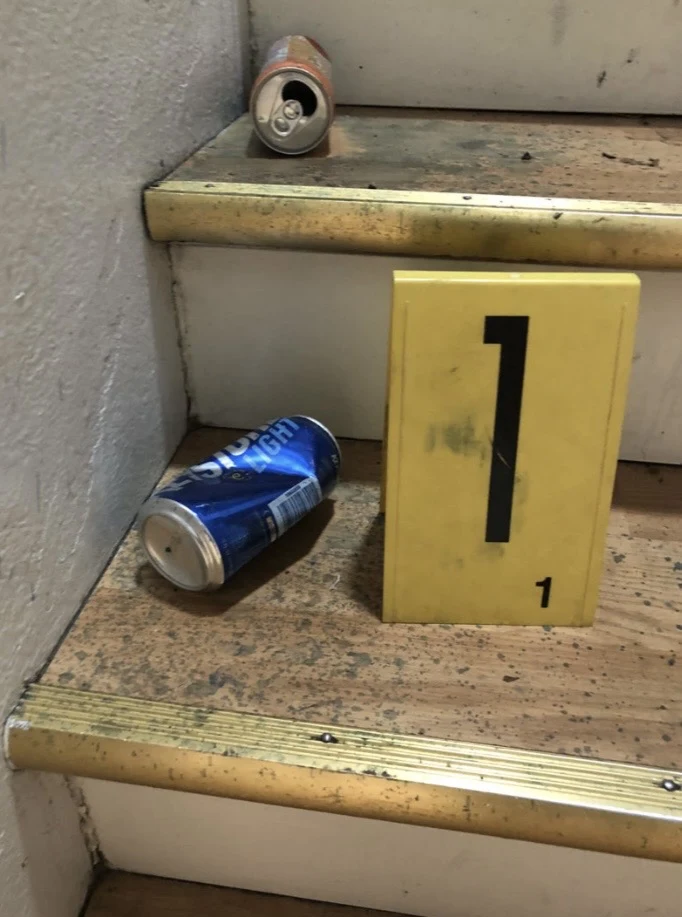 Blue beverage can on a worn stair tread beside yellow evidence marker 1, with another can on the step above.