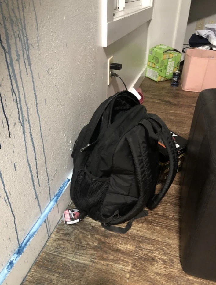 Black backpack on a wooden floor beside a wall with blue drip marks, with boxes and a pink container behind it.