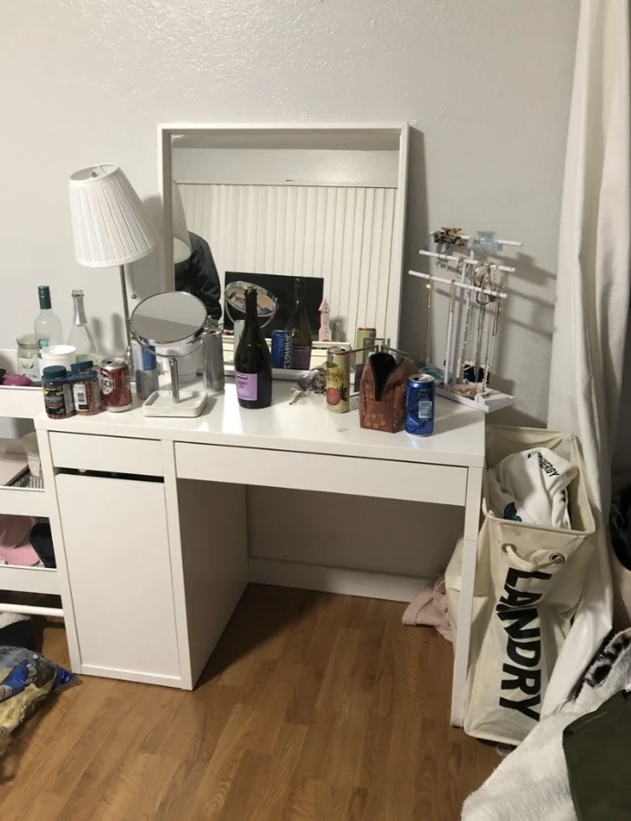 White bedroom vanity desk with mirror, lamp, jewelry stand, bottles and cans, beside a fabric laundry hamper.