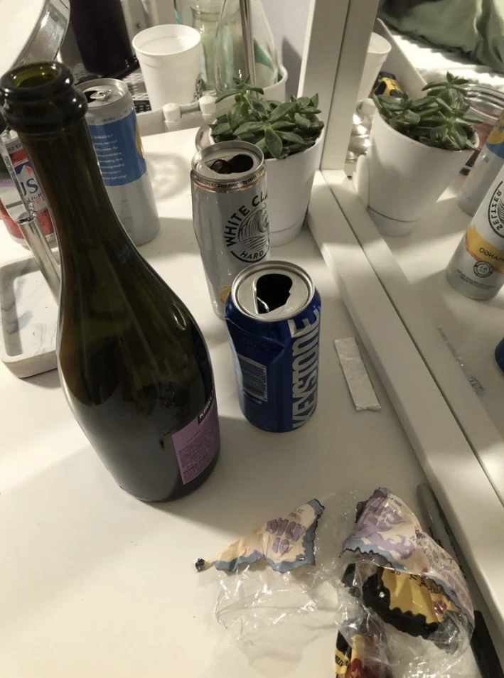 Vanity surface and mirror with bottles, open beverage cans, a small potted succulent and wrappers.