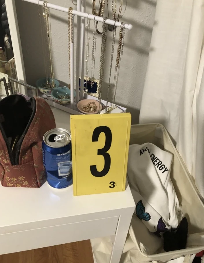 Blue beverage can, jewelry stand and pouch on a white vanity beside yellow evidence marker 3 and a laundry hamper.