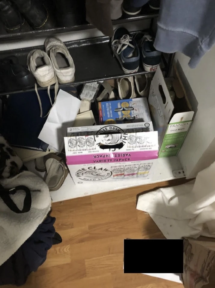 Closet floor with a shoe rack, shoes, beverage cartons and loose clothing, with a small black redaction.
