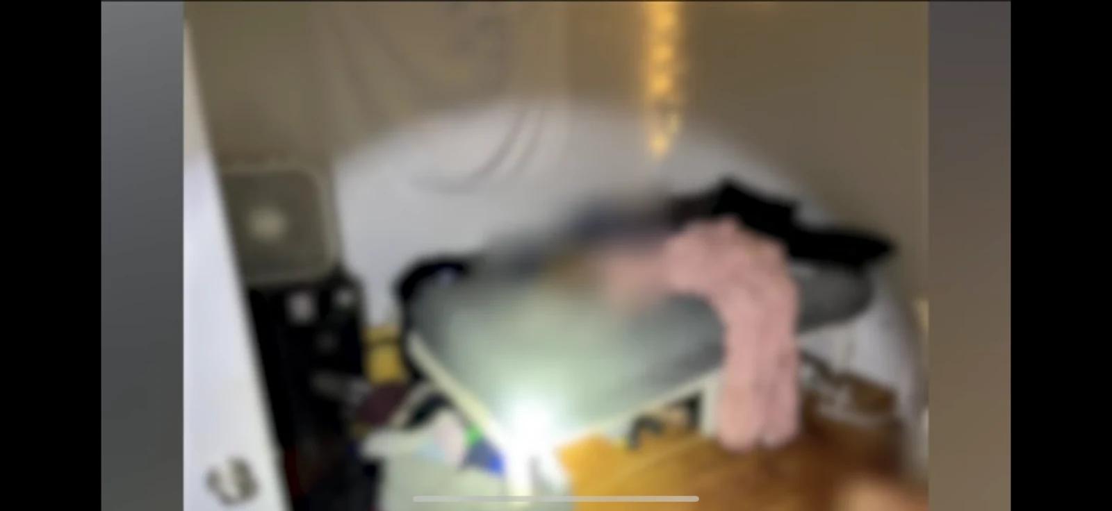 Soft-focus view through a bedroom doorway toward a bed with gray bedding, a pink blanket and scattered items.