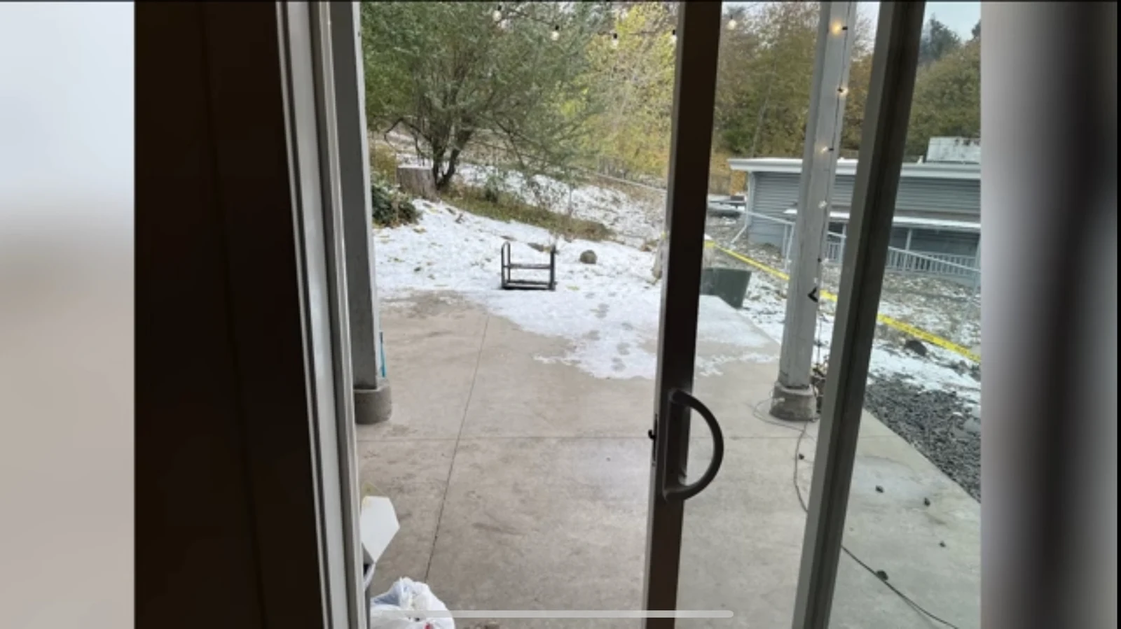 View through a sliding glass door onto a concrete patio, snow-covered ground, trees and outdoor posts.