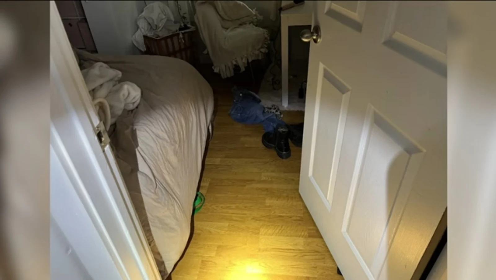 View through a bedroom doorway showing a bed, wooden floor, clothing, shoes and a laundry basket.