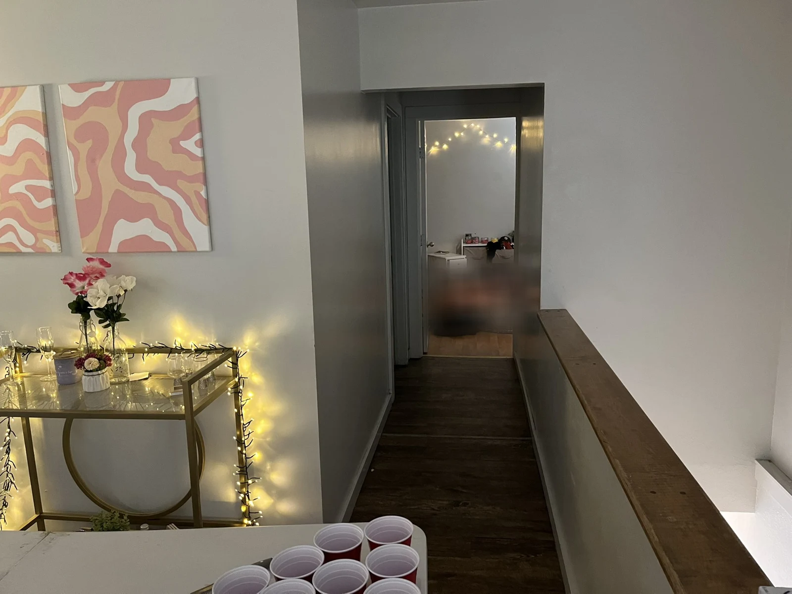 Hallway beside a glass console with string lights and flowers, abstract wall art and red cups on a table, with a blurred area in the distant room.