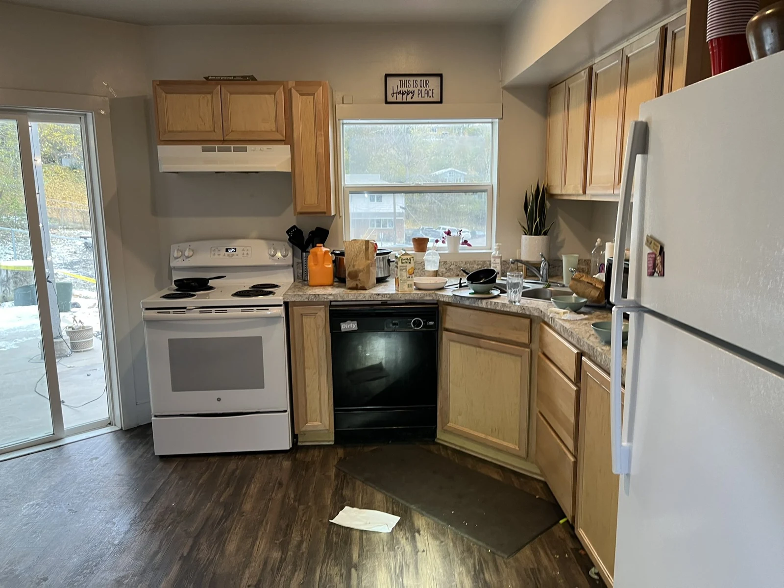 Kitchen with wooden cabinets, white stove and refrigerator, black dishwasher, sink, countertop items and a sliding glass door.