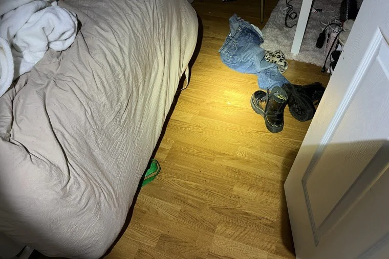 Bedroom floor beside a bed, with jeans, shoes and a flashlight-lit patch of wood flooring.