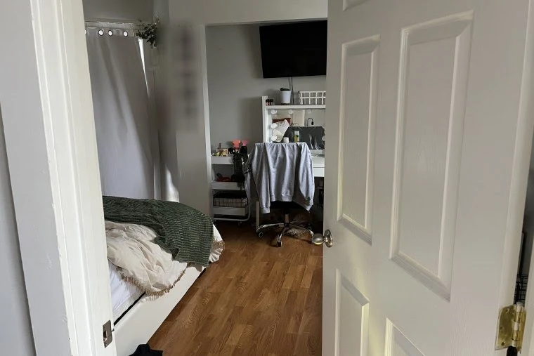 Bedroom doorway showing a bed with green and white bedding, a desk and chair, shelves and a wall-mounted television.
