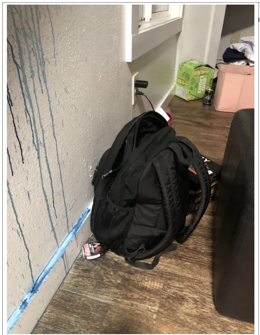 Black backpack beside a wall with blue drips, on a wooden floor near boxes and a pink container.